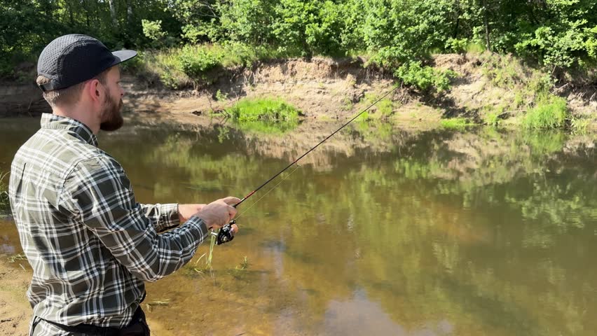 A bearded young man with a spinning rod catches fish on the river bank and smiles. Fisherman on the weekend. Hobbies, recreation and active recreation in summer