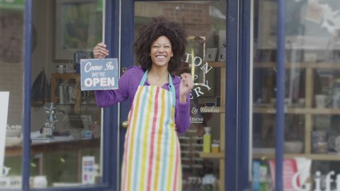 A happy and excited female shopkeeper stands outside of her small independent shop to welcome potential customers. She is holding an 'open' sign and wearing a colorful apron.