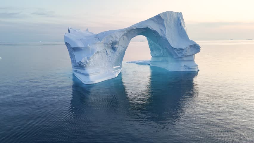 A tall melting iceberg in the Arctic Ocean. A glacier with a huge hole resembling an ice gate. Clear ice floats in turquoise water. Ecology, melting ice, climate change, global warming. Aerial view. M
