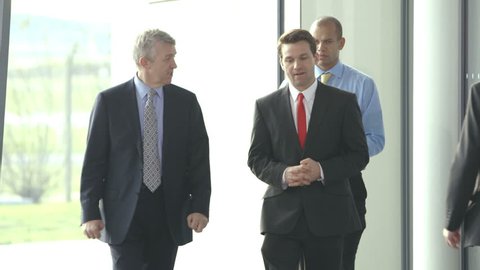 Two businessmen chat together as they walk along through a busy modern office building.