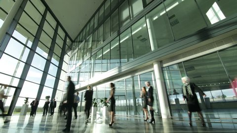 Time lapse of a large group business people moving around a large, open plan, glass fronted office building. The interior panels of glass are reflecting the clouds outside as they move across the sky.