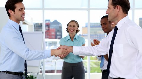Side view of smiling Caucasian businessmen shaking hands while other multi-ethnic business people applauding in bright office