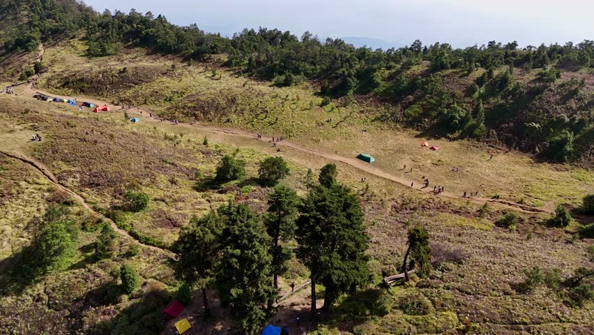 Drone footage showing crowded hiking path cutting through dry grassy hills, surrounded by forest trees under clear blue skies. Mount Prau, Indonesia.