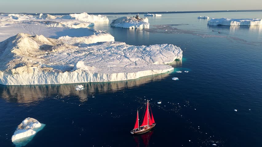 Little red sailboat cruising among floating icebergs in Disko Bay glacier during midnight sun season of polar summer. Ilulissat, Greenland. Global warming and melting glaciers.