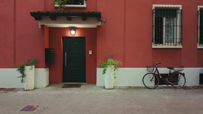 Static shot of a red building entrance in Brescia with a dark green door, two tall white planters, barred windows, and a vintage bicycle parked nearby during daytime.