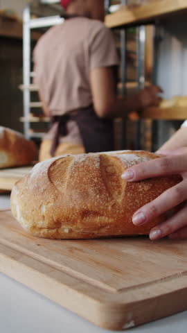Vertical close-up view of hands of unrecognisable woman using serrated knife to slice round loaf of white bread on wooden cutting board while female baker working in blurred background