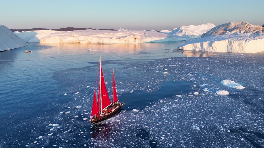 Little red sailboat cruising among floating icebergs in Disko Bay glacier during midnight sun season of polar summer. Ilulissat, Greenland. Global warming and melting glaciers.