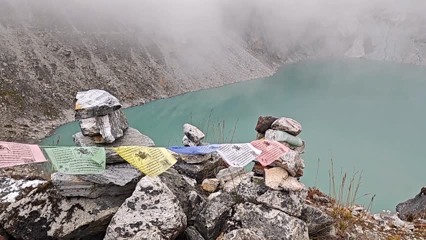 Scenic mountain view featuring vibrant prayer flags draped over carefully arranged stones near a calm turquoise lake, surrounded by mist and rocky terrain, evoking tranquility and harmony