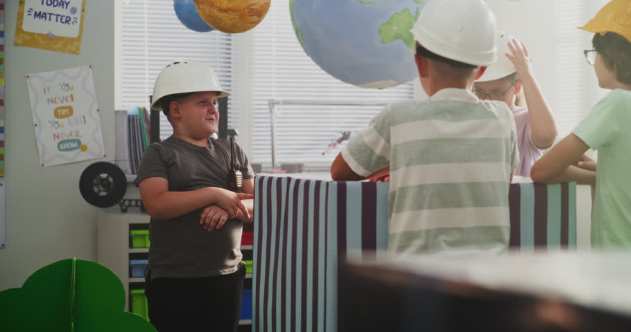 Team of Primary School Students in Hardhats Discussing City Planning, Young Boy Giving Instructions. Interactive STEM Class with Large Cardboard Urban Blocks and Models of Trees, Buildings. Dolly Shot