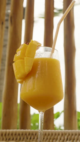 Full glass of mango smoothie with large bamboo straw and mango garnish on wicker table with palm trees behind, mid shot, Tropical Vacation Concept.