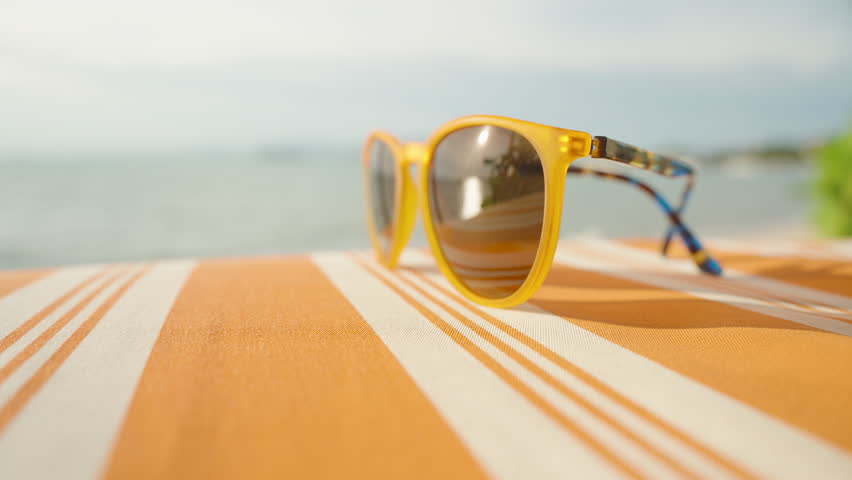 Yellow sunglasses placed on striped orange sunbed with ocean view in background, bright lighting and reflection, Summer Vibe Concept.