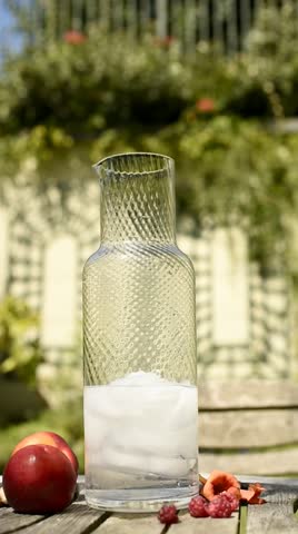 Fresh, clear water is poured into a tall glass jug with ice. Juicy nectarines rest beside it on an outdoor table in a summer garden on a hot day. Vertical video. FullHD.