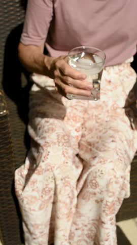 A close-up shot of a senior woman sitting on a sunny terrace, enjoying a refreshing glass of water with ice. The video emphasizes the importance of hydration on a hot summer day. Vertical, FullHD.
