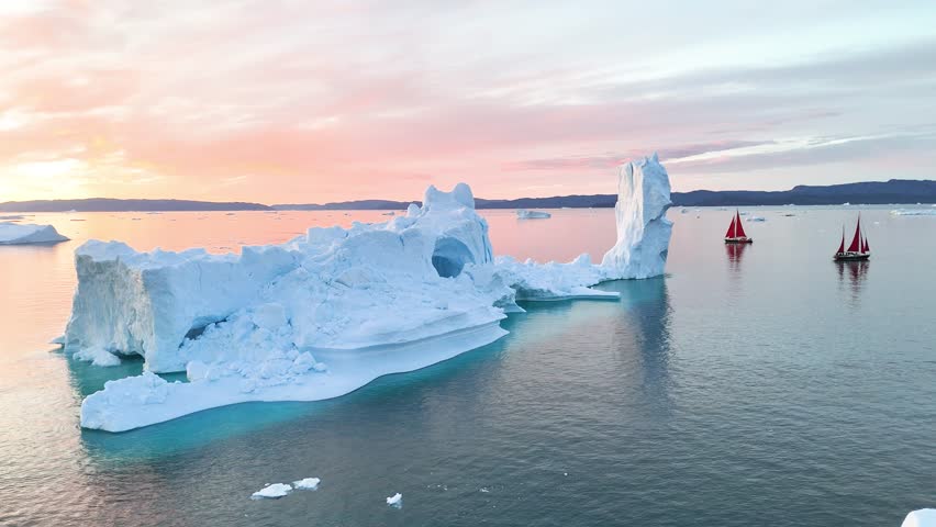 Little red sailboat cruising among floating icebergs in Disko Bay glacier during midnight sun season of polar summer. Ilulissat, Greenland. Global warming and melting glaciers.