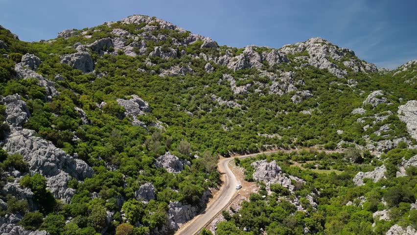 Drone spirals above car in Turkish highlands. Rocky hills and green forest frame road. Turkiye terrain seen from air. Remote road trip beauty in Turkey