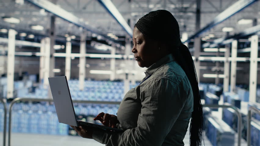 IT expert monitors server farm activity using laptop while walking in facility. African american woman in data center reviews rigs tests results to ensure systems are working correctly, camera B