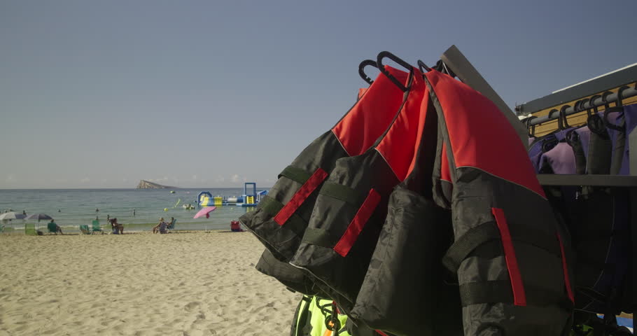 Bright orange life jackets hang on a rack by the beach, with people relaxing on the sand and an inflatable water park floating in the sea in the background. A sunny coastal holiday scene.