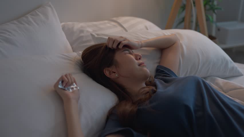 Depressed Asian woman feel upset while lying down on bed in bedroom. Frustrated stressed girl feeling sad and headache with life problem and financial in house and hold pack of medicine capsule pill.