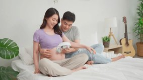 Asian expectant couple sharing loving moment, ready to become parents. Attractive pregnant wife and husband placing headphones on belly to play music for baby while sitting on cozy bed in bedroom. - Powered by Shutterstock - Get 15% off with code: PIKWIZARD15
