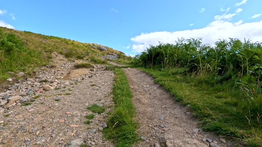 A steady walk up a rugged, rocky hillside trail bordered by grass and shrubs under bright daylight, with smooth handheld camera movement and wide landscape views