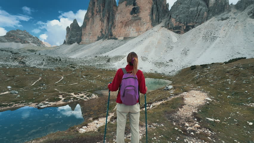 Lone female hiker walking remote alpine trail, approaching iconic Tre Cime di Lavaredo peaks under bright summer sky in scenic Dolomites mountain landscape