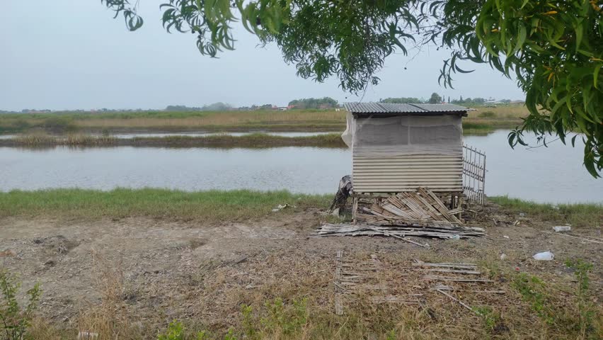 A small, weathered hut stands by the calm riverside, surrounded by dry grass and debris, while nearby tree leaves sway gently in the breeze under an overcast sky.