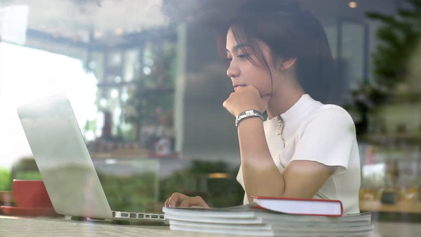 Portrait from a window of an Asian teenage girl working on a laptop in a cozy coffee shop on a bright sunny day, enjoying a relaxed atmosphere with warm natural light streaming into the café