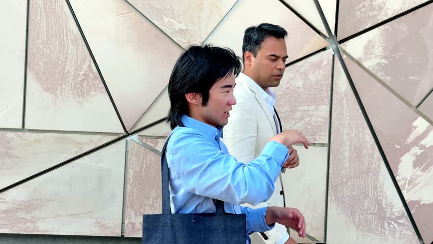 Two businessmen, one Asian and one Indian, walk and talk together near Federation Square in Melbourne, Australia, discussing business matters during a coffee break