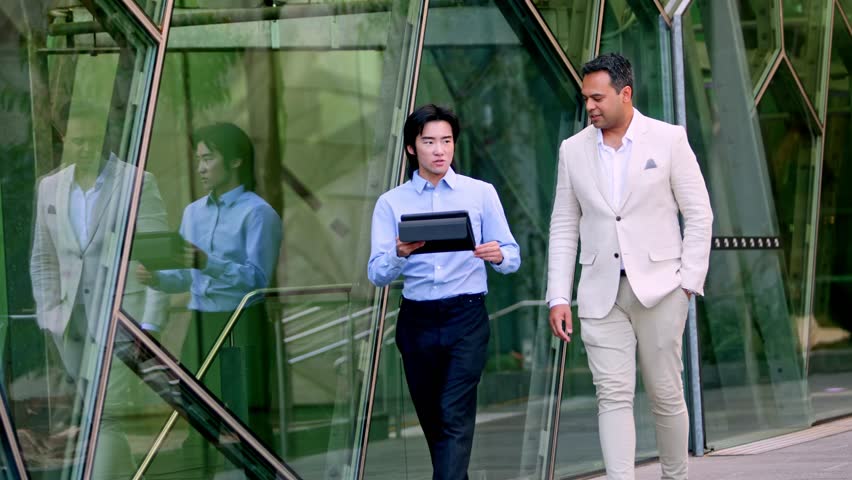 Two businessmen, one indian and one chinese, are walking and talking in front of a modern glass building in Melbourne, Australia, while the chinese businessman holds a tablet