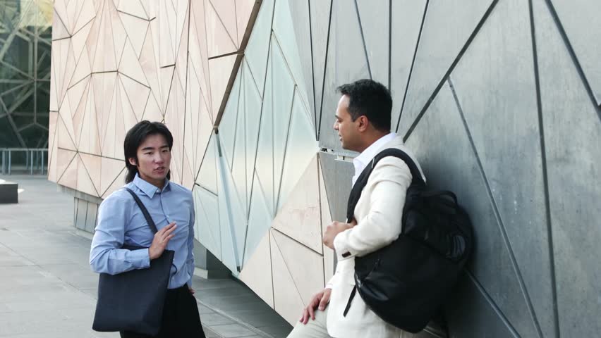 Business partners from India and China engaging in professional discussion during coffee break near Federation Square, exchanging ideas in urban Melbourne setting with modern architectural backdrop