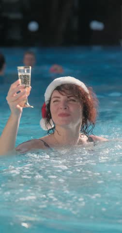 Woman in Santa hat enjoying champagne while standing in a swimming pool, representing tropical Christmas and festive holiday celebration