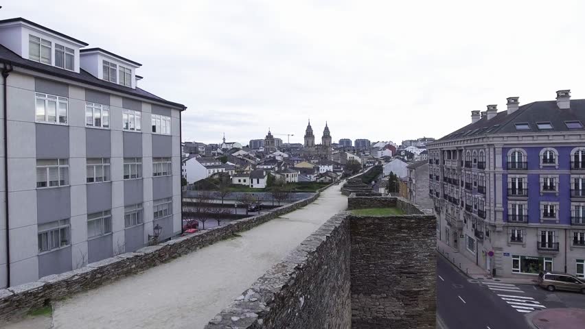 Aerial drone takeoff from Lugo’s Roman wall walkway revealing the Cathedral and the surrounding ring road. People and vehicles visible. UNESCO World Heritage Site.