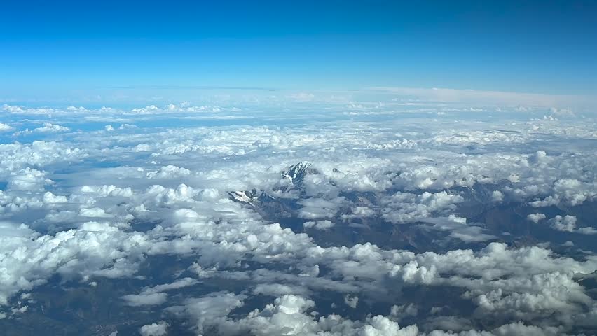 An elevated aerial view of the Monta Blanc summit taken from a jet cockpit, with the mountaintop with almost no snoy in. a sumer day, surroended by cottony clouds under a blue and bright sky.