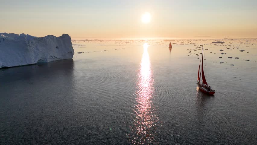 Little red sailboat cruising among floating icebergs in Disko Bay glacier during midnight sun season of polar summer. Ilulissat, Greenland. Global warming and melting glaciers.