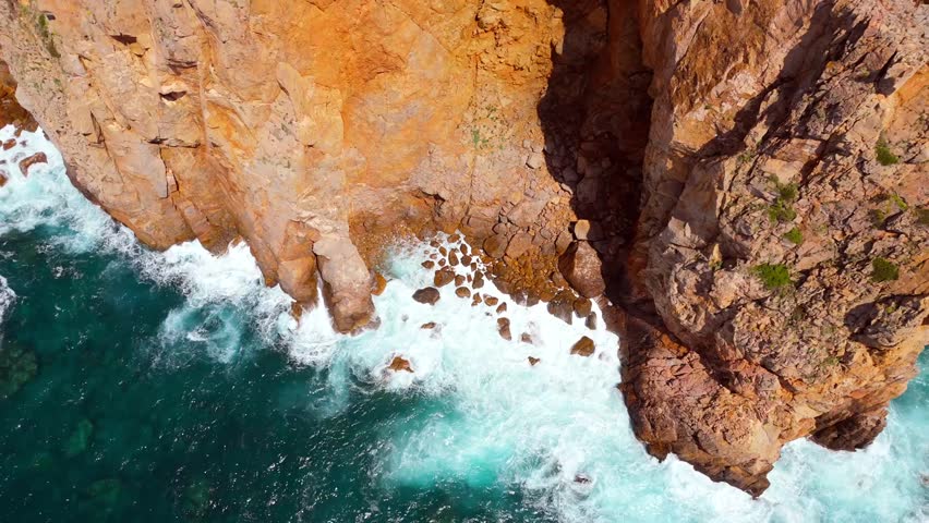 Aerial view of rugged cliffs in Sardinia, Italy, with powerful ocean waves crashing against the rocks, showcasing dramatic coastal scenery.