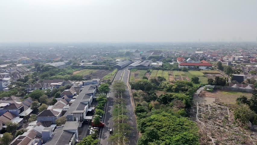 An aerial shot shows a quiet residential area with a tree-lined main road. The camera slowly pans to the right, revealing more houses and green spaces.