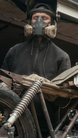 Vertical low angle shot of unrecognizable man in protective helmet and gloves using welding machine on his bike in garage