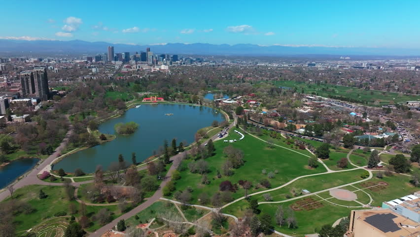 City Park Denver Colorado Lake Pavilion vibrant spring summer aerial drone sunny blue sky snow capped Rocky Mountains front range cityscape green lush grass trees blossom pan left motion