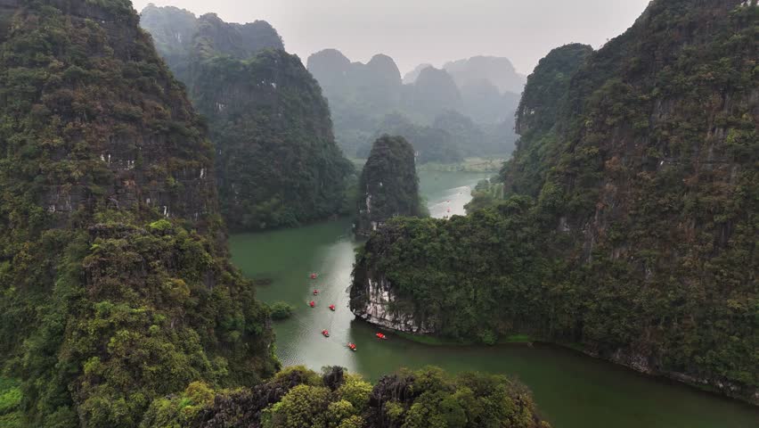 Traditional sampan boats navigate the winding Sao Khe River between towering limestone karsts in Trang An, Ninh Binh, Vietnam, captured by ascending drone shot.