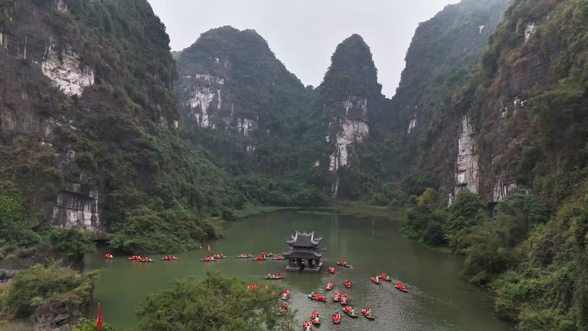 Sampan boats surround a traditional riverside pavilion set between towering limestone cliffs in the UNESCO-listed Trang An Landscape Complex, Ninh Binh, Vietnam, captured by aerial drone shot.