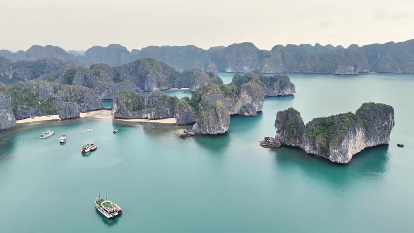 Tourist boats anchor beside secluded sandy beaches and towering limestone karsts in UNESCO-listed Ha Long Bay, Quang Ninh Province, Vietnam, captured by orbiting aerial drone shot.