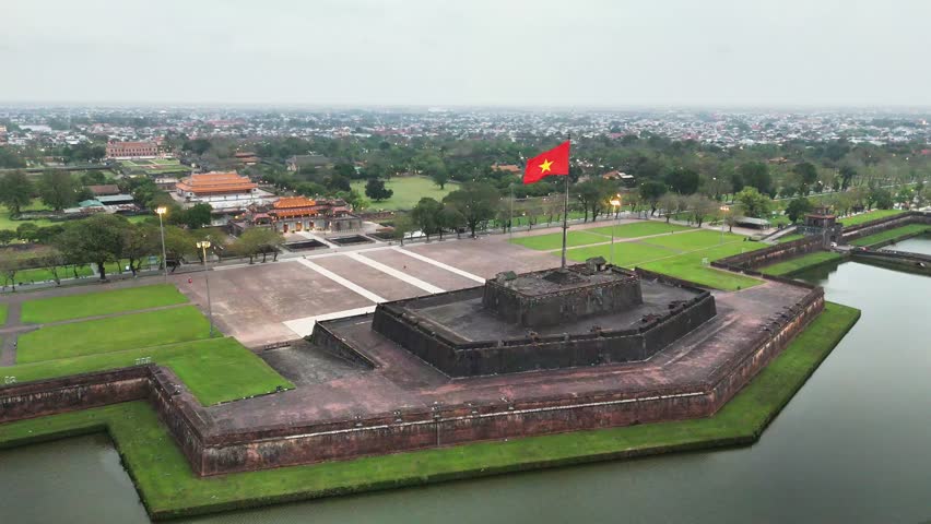 Large Vietnamese flag waves atop the historic flag tower of the UNESCO-listed Hue Citadel, with the Ngo Mon Gate and Imperial City beyond, central Vietnam, captured by static drone shot.