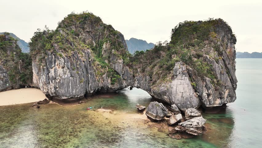 Dramatic limestone cliffs connected by a natural rock arch rise above a sandy beach and shallow turquoise waters in UNESCO-listed Ha Long Bay, Quang Ninh Province, Vietnam, captured by pull-out drone.