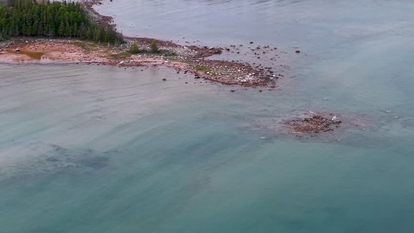 Aerial drone view of rocky Lake Huron shoreline with shallow turquoise waters and scattered boulders near forested coast in Michigan’s Upper Peninsula
