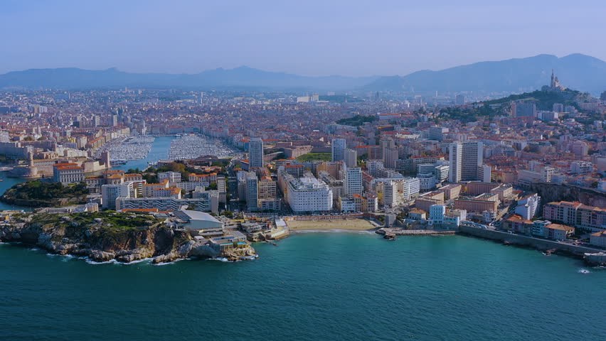 4K drone panorama of Marseille : Vieux-Port packed with yachts, Catalans Beach and Palais du Pharo at the entrance, Notre-Dame de la Garde on the hill, Mediterranean coast and Provence hills beyond.