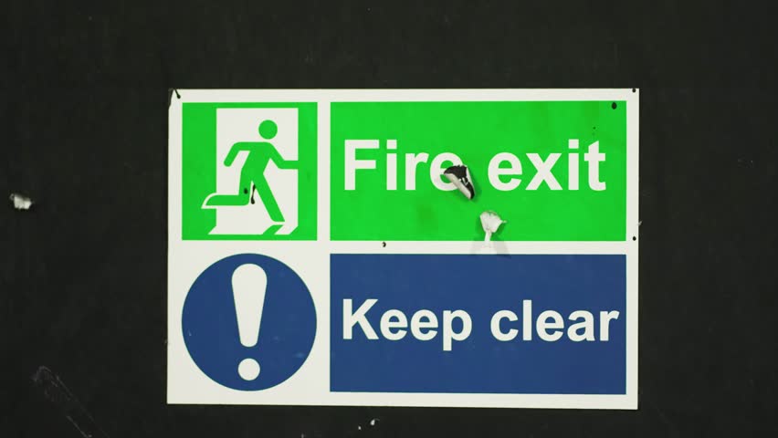 Sign combining fire exit and keep clear warnings mounted on dark wall. Board displaying instruction with text fire exit keep clear and emergency symbol. Sticker emphasizing escape route visibility in