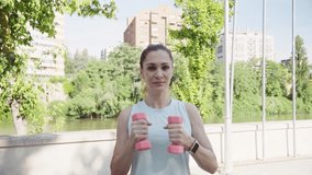 Fitness instructor is demonstrating alternating bicep curls using light pink dumbbells, focusing on proper form and controlled movements in a park setting near a river - Powered by Shutterstock - Get 15% off with code: PIKWIZARD15