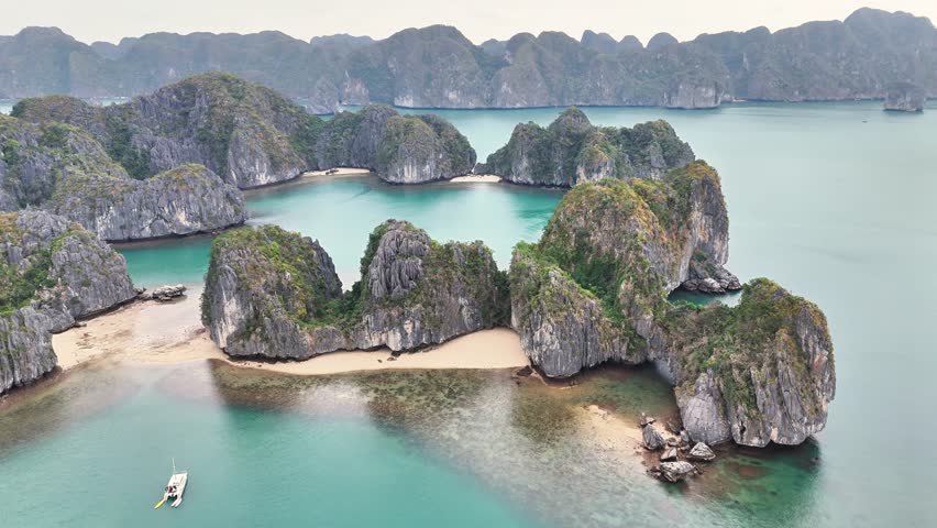 Aerial view over Ha Long Bay, Vietnam, featuring limestone karsts, sandy beaches, and turquoise waters, with a boat anchored nearby amidst the stunning seascape and scattered lush green islets
