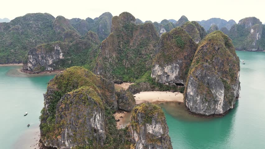 Aerial view of Ha Long Bay, Vietnam, featuring towering limestone islands, secluded sandy beaches, and turquoise waters surrounded by lush green hills under a calm, overcast sky