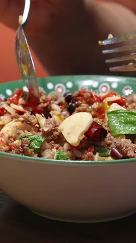 A person uses a spoon and fork to scoop up spicy beef, garlic, chili, and rice from a colorful bowl under warm indoor lighting, close-up view